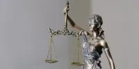A metal statue of Lady Justice, blindfolded, holding a set of balanced scales in her raised hand against a plain grey background
